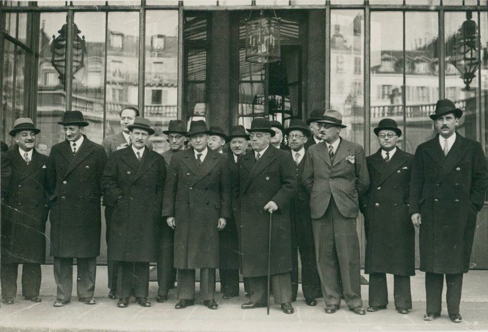 photo noir et blanc : une quinzaine d'hommes posent debout, en costome, manteau et chapeau, devant une vitrine