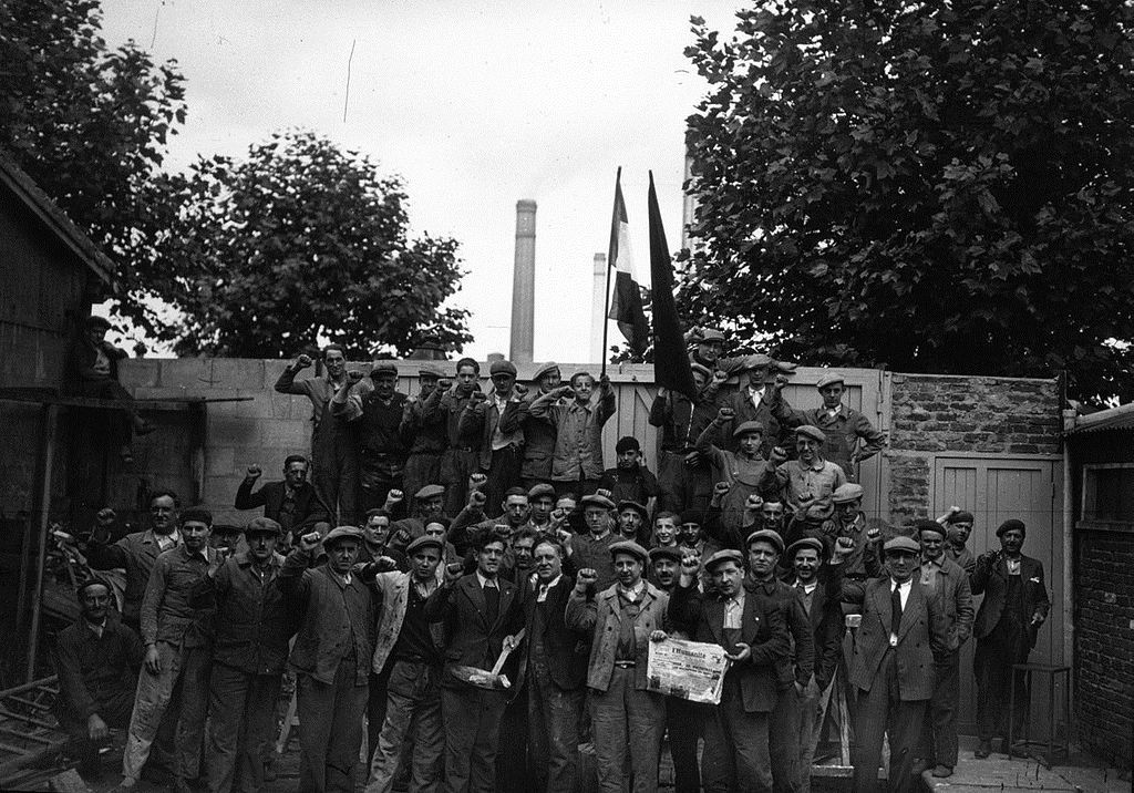 photo noir et blanc d'une foule d'ouvriers faisant corps devant l'entrée d'une usine en levant le poing
